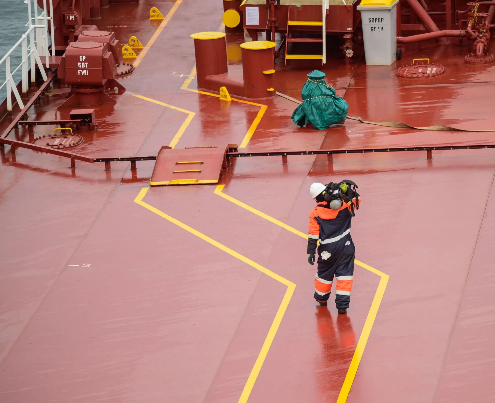 A worker in a hard hat and high-visibility overalls walking across the red deck of a ship