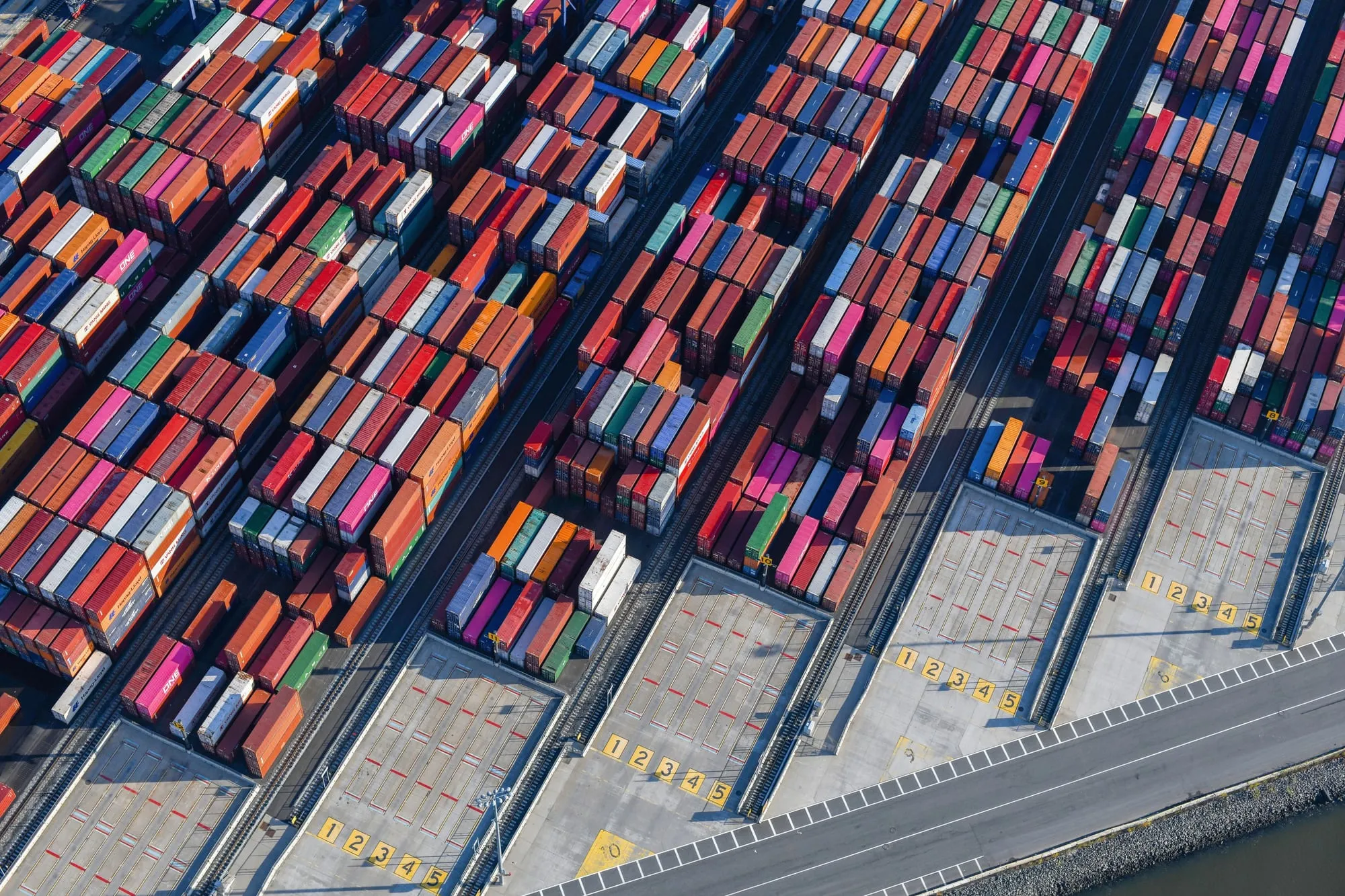 Aerial view of a container terminal: rows of stacked shipping containers beside numbered loading bays on the quay